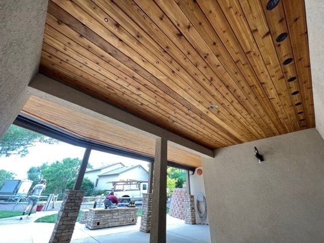 Wooden plank ceiling over an outdoor patio space with natural light and recessed lights.  View of a street scene visible through open space.
