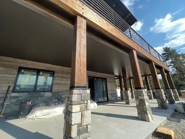 A covered patio with wooden columns and stone bases supports a second-story deck with a metal railing.