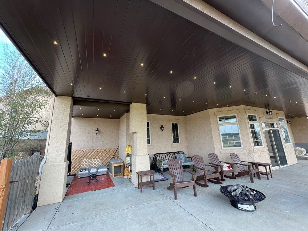 Patio with brown ceiling and recessed lights, furnished with seating and fire pit. Beige stucco building with windows and door.