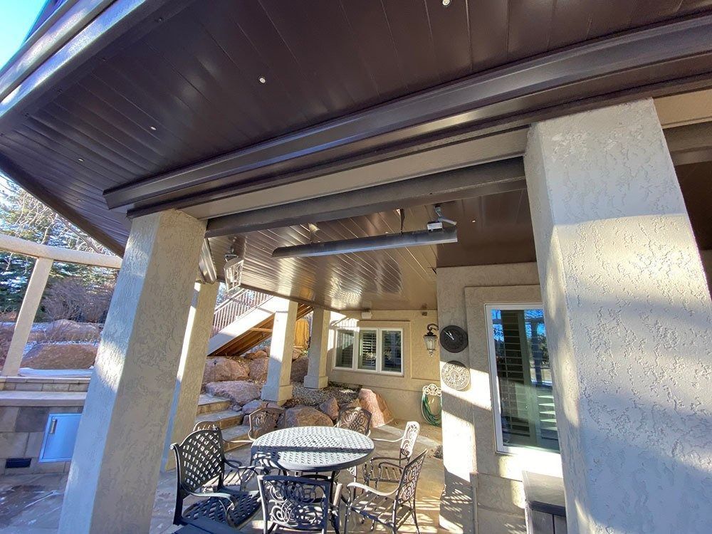 Patio with brown awning and pillars, a table, chairs, and a wall heater. Sunlight illuminates the space.