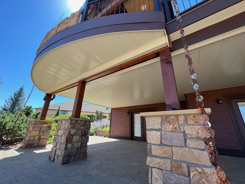 Low-angle view of a deck with brown and beige trim, supported by stone-covered columns. A rain chain hangs down.