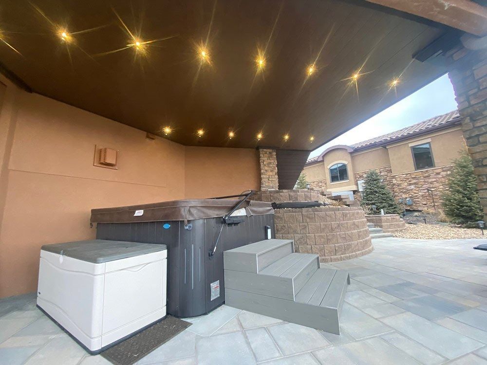 A covered outdoor hot tub area with a hot tub, storage box, and gray steps. Warm lighting illuminates the ceiling and a stone wall.