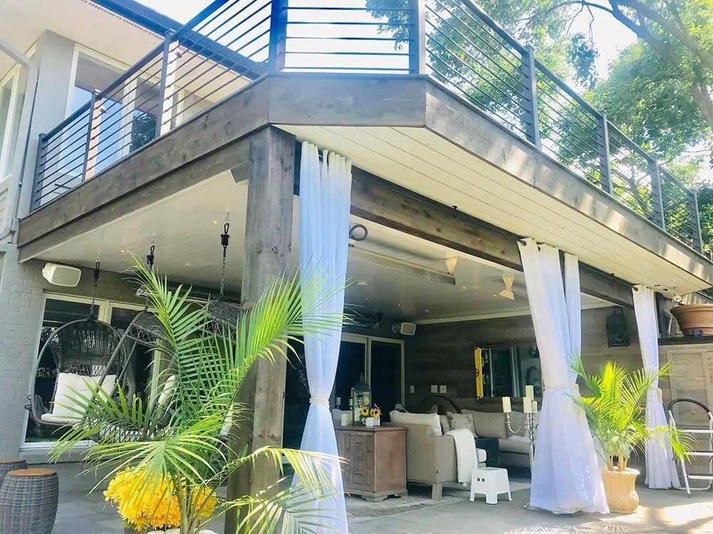 Deck with white curtains, a seating area below, and greenery.  The deck has metal railings and is supported by wooden beams.