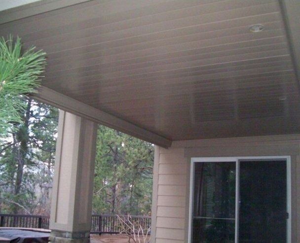 Beige porch ceiling with horizontal planks, a pillar, and a glass sliding door overlooking trees.