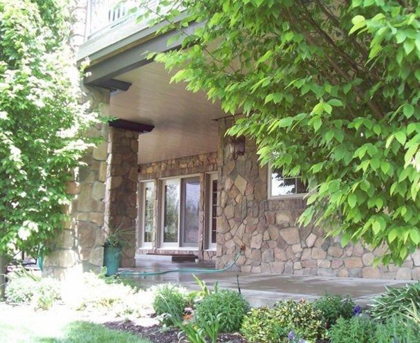 Stone-walled house with a porch and columns, partially obscured by green trees. Sunlight streams onto the porch.
