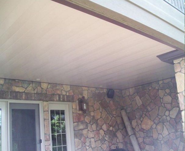 Stone-walled porch ceiling with wood-look paneling. A window and door are on the left side, a speaker and rolled-up rug on the right.