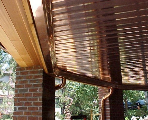 Copper gutters and downspouts installed on a brick and wood-framed porch. The metal reflects the warm sunlight.