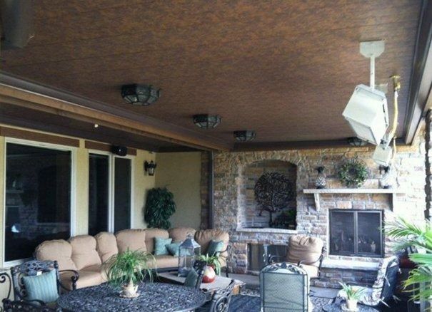Outdoor patio with a stone fireplace, couches, and ceiling-mounted heaters.  Brown ceiling and stone wall, with windows and lush greenery.