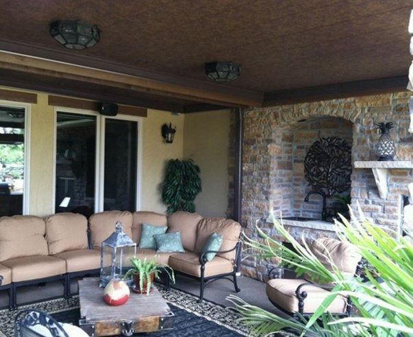 Outdoor patio with beige couches, a stone fireplace, and plants, under a brown ceiling with two light fixtures.