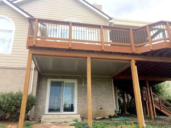 A two-story wooden deck attached to a house with a sliding glass door below. Brown railings and support beams, with a beige ceiling under the deck.