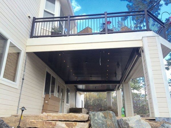 Beige house with a black-railed deck, overlooking a covered patio supported by white columns, with rock landscaping.