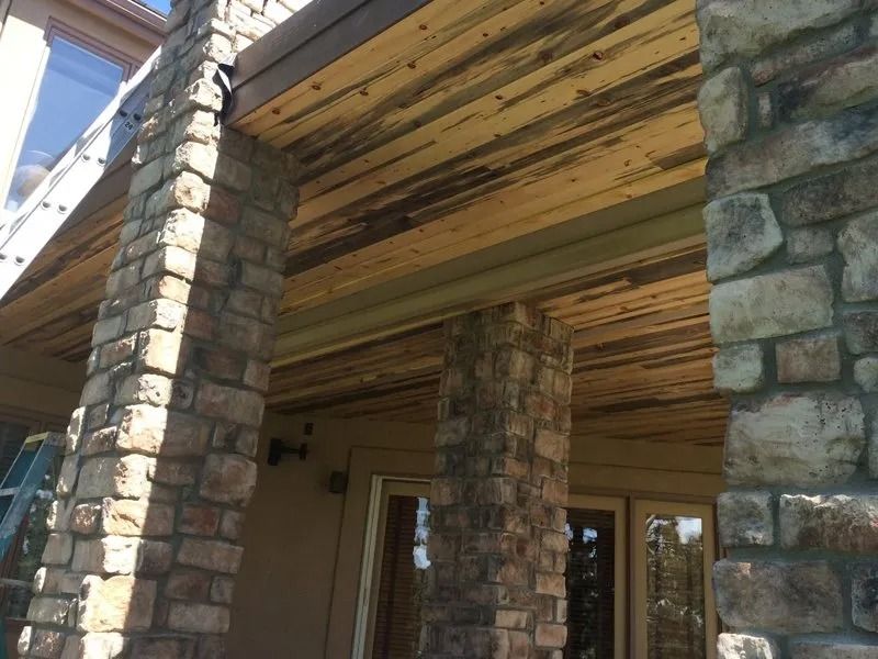 Stone pillars support a wooden porch ceiling with weathered, stained wood. The setting appears to be a home's exterior.