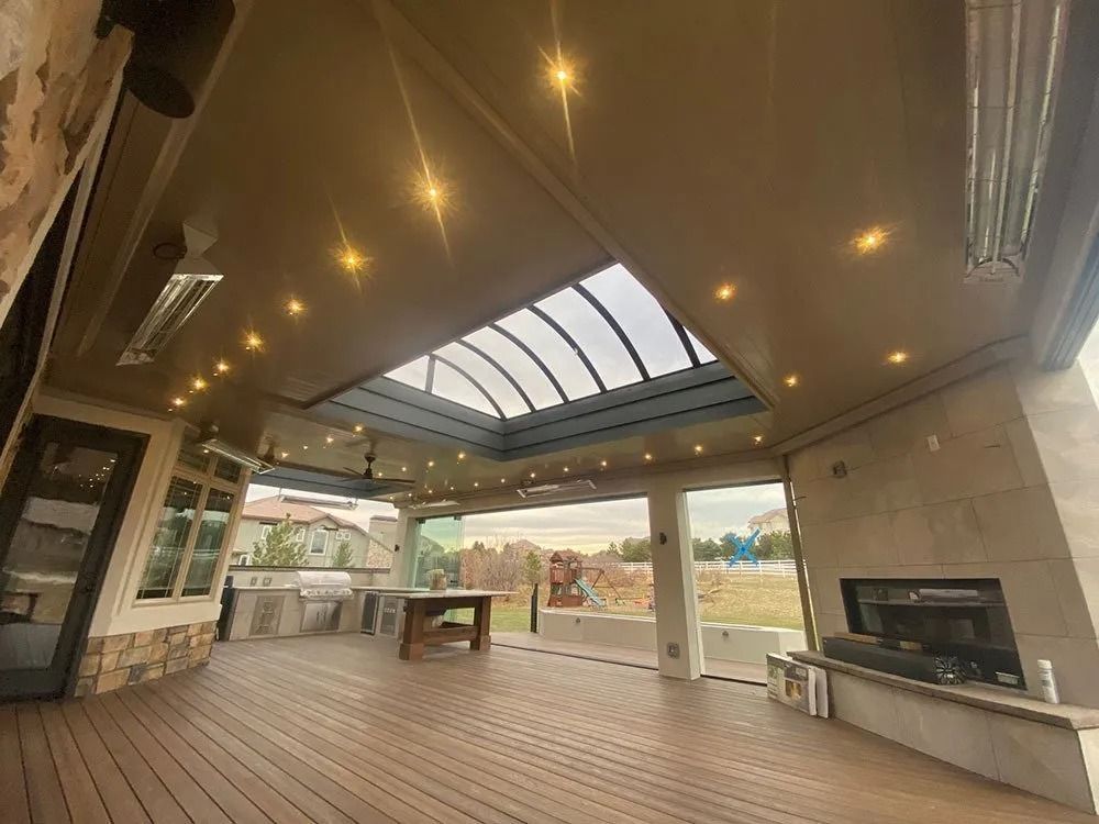 A covered outdoor patio with wood flooring, a fireplace, and a retractable roof panel. It has recessed lighting and heating elements.