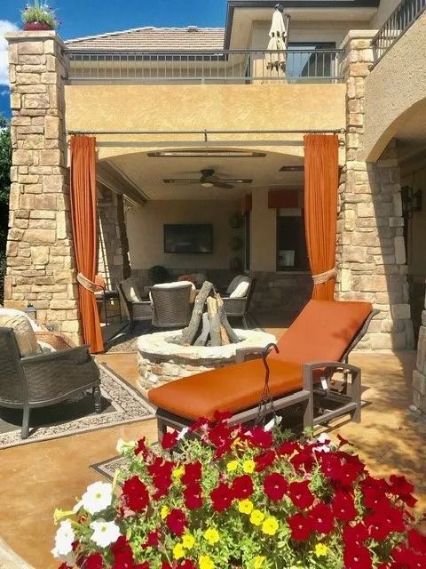 Outdoor patio with a fire pit, lounge chair, and curtains, surrounded by flowers and stone columns.