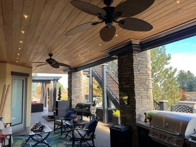 Outdoor patio with wood ceiling, fans, and stone pillars. Seating, grill, and views of surrounding trees are also visible.