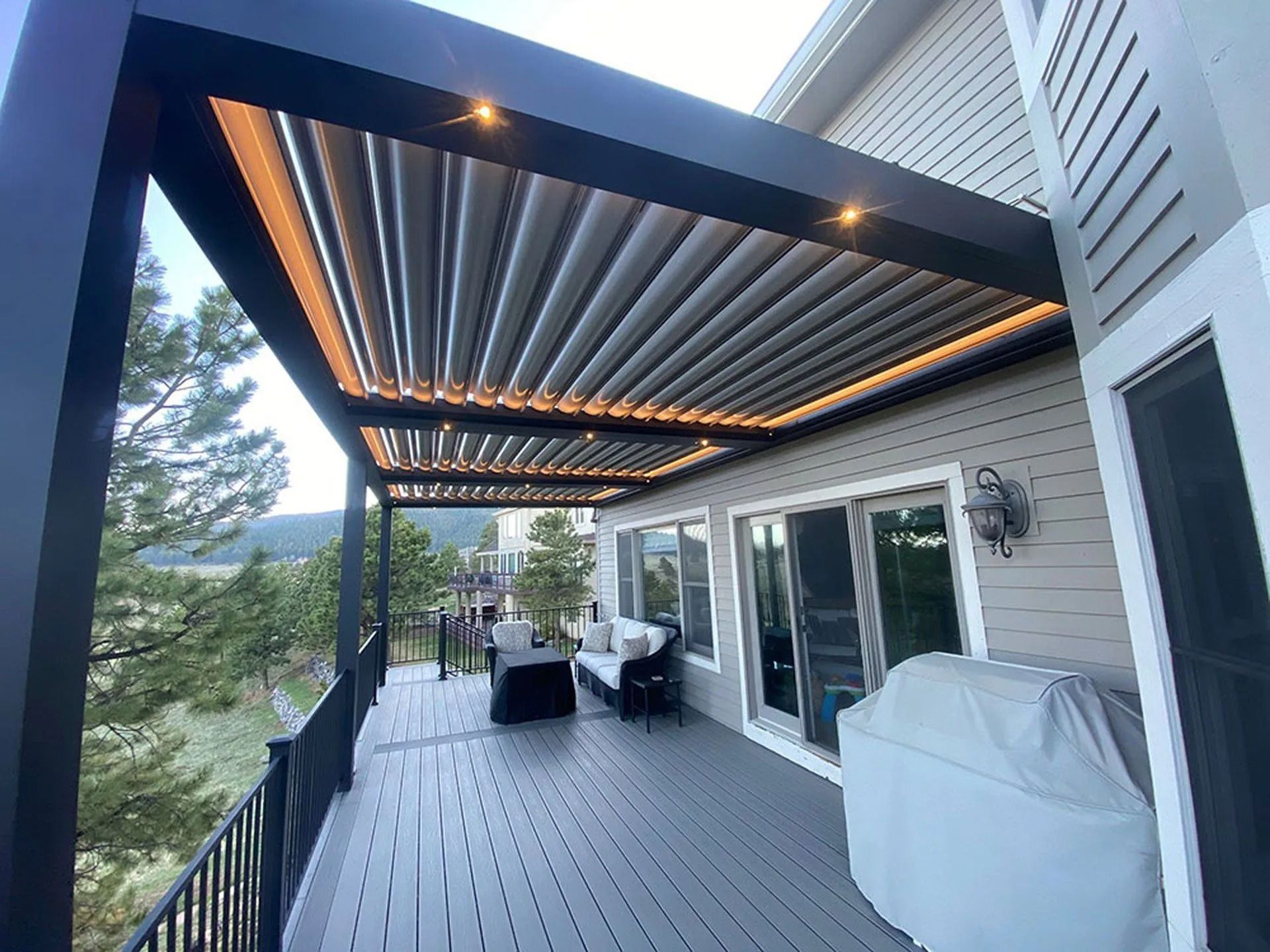 A modern deck with a black pergola, built-in lights, and retractable roof; overlooks a scenic view with a grill.