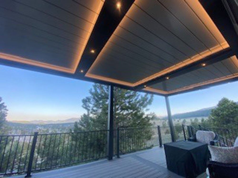 Outdoor patio with a modern, dark gray, illuminated pergola overlooking a scenic view of trees and mountains.