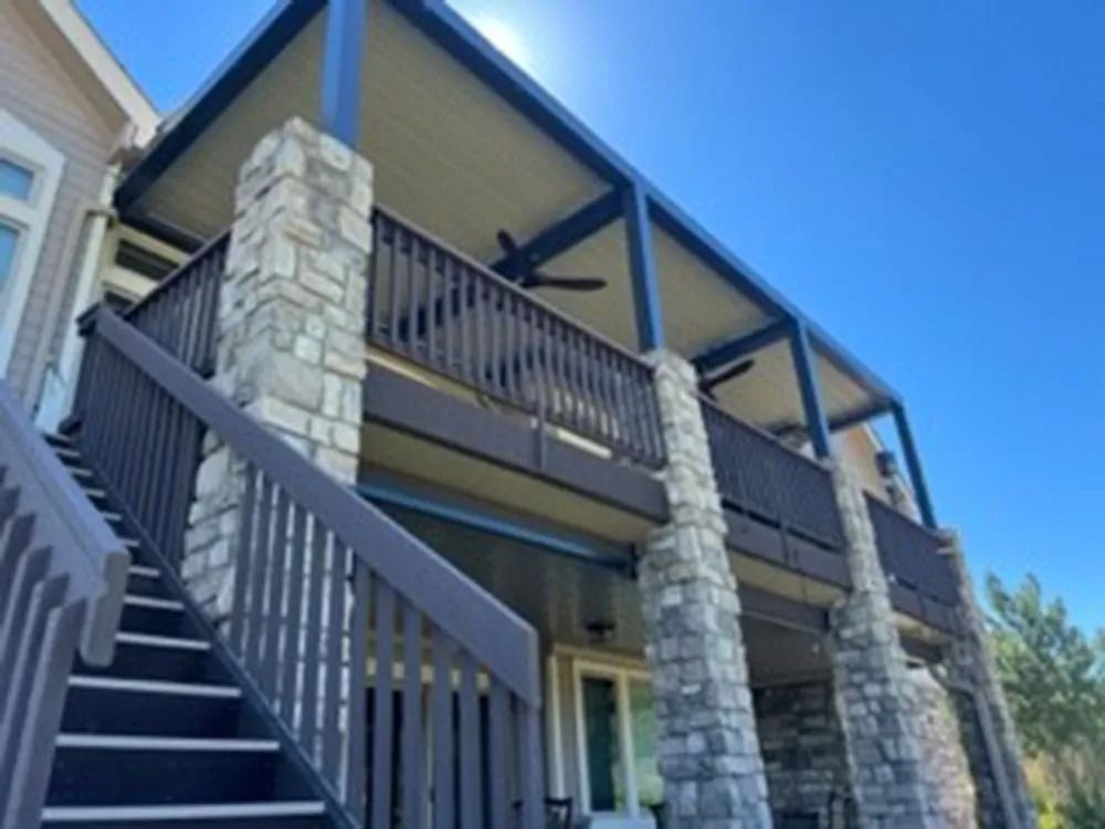Deck with stone columns and dark wood railings, topped with a covered patio in the sun.