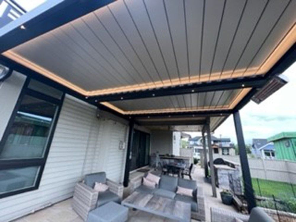 A modern outdoor patio with a black pergola, beige ceiling, and built-in lighting. Patio furniture includes a sofa, chairs, and a dining table.