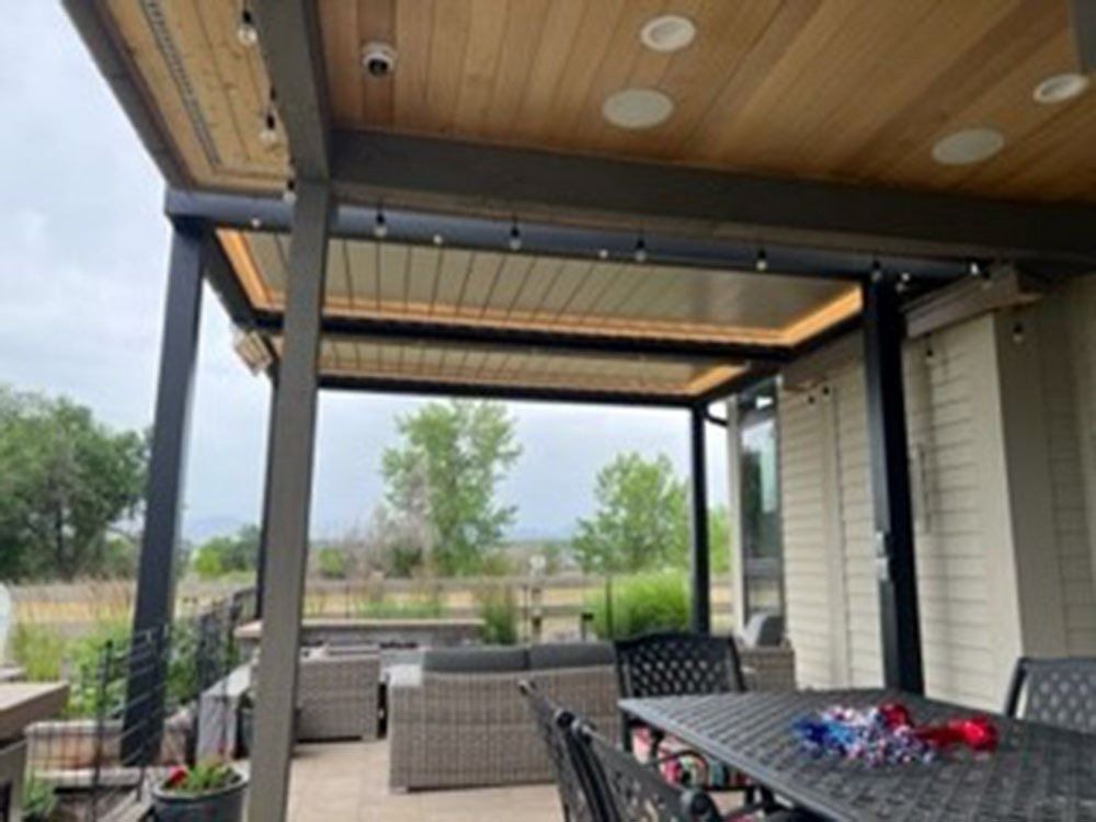 Patio with retractable roof, wooden ceiling, string lights, and outdoor furniture. Cloudy day with view of trees and landscape.