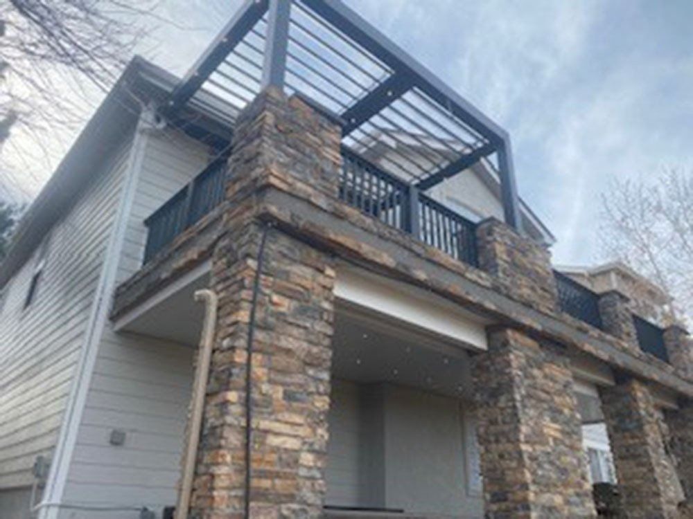 A two-story house with a stone-covered porch and a black pergola on the roof. The house has tan siding and a cloudy sky.