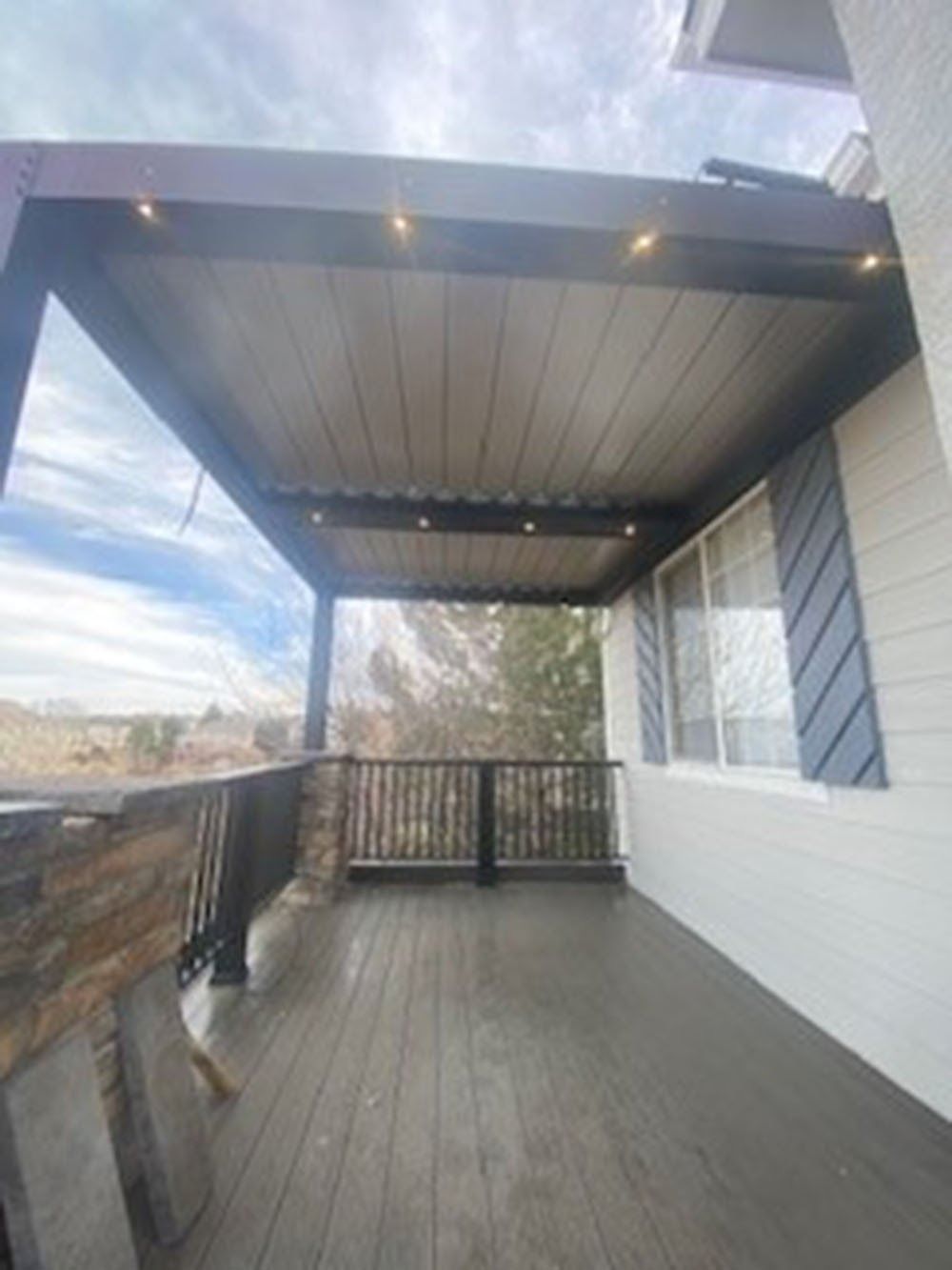 Covered deck with gray flooring, black railing, and string lights, overlooking a yard. The structure is connected to a white house with blue shutters.