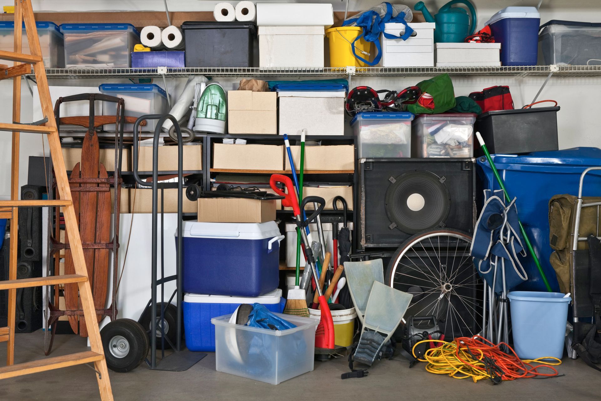 Cluttered garage filled with storage bins, tools, cleaning supplies, and household items stacked on shelves and the floor.