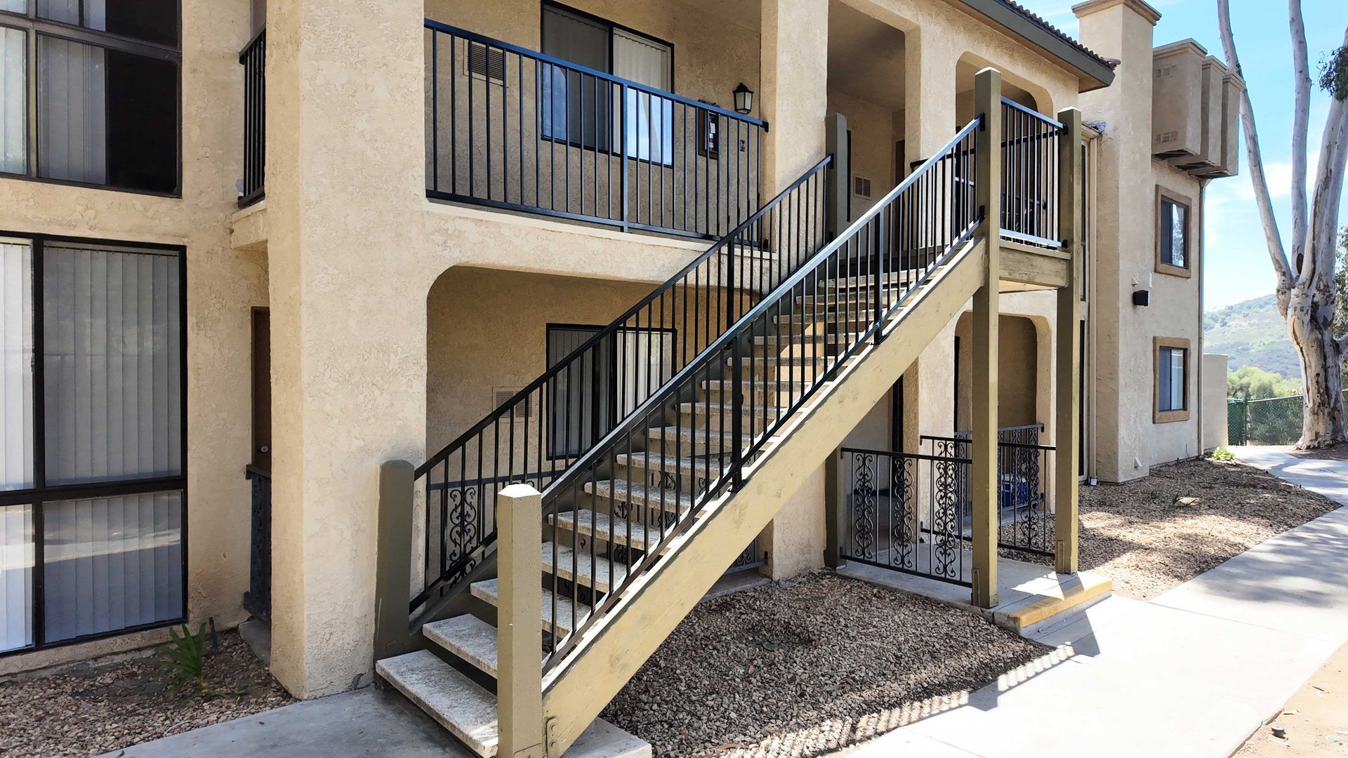 Apartment building exterior with staircase leading to second-floor units. Beige stucco, black railings, sunny day.