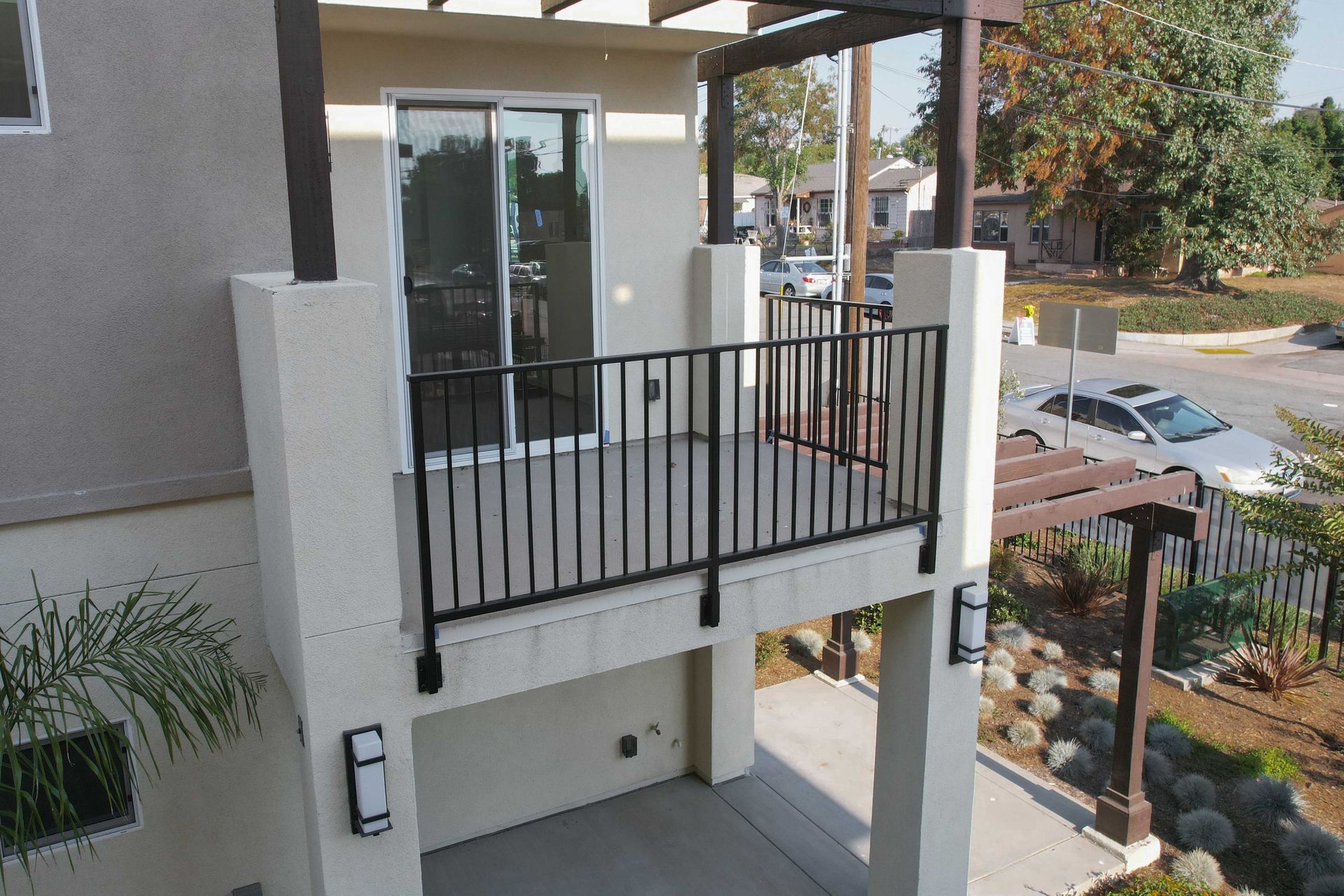 Balcony with black railing, sliding glass door, and wooden pergola, overlooking a parking lot.