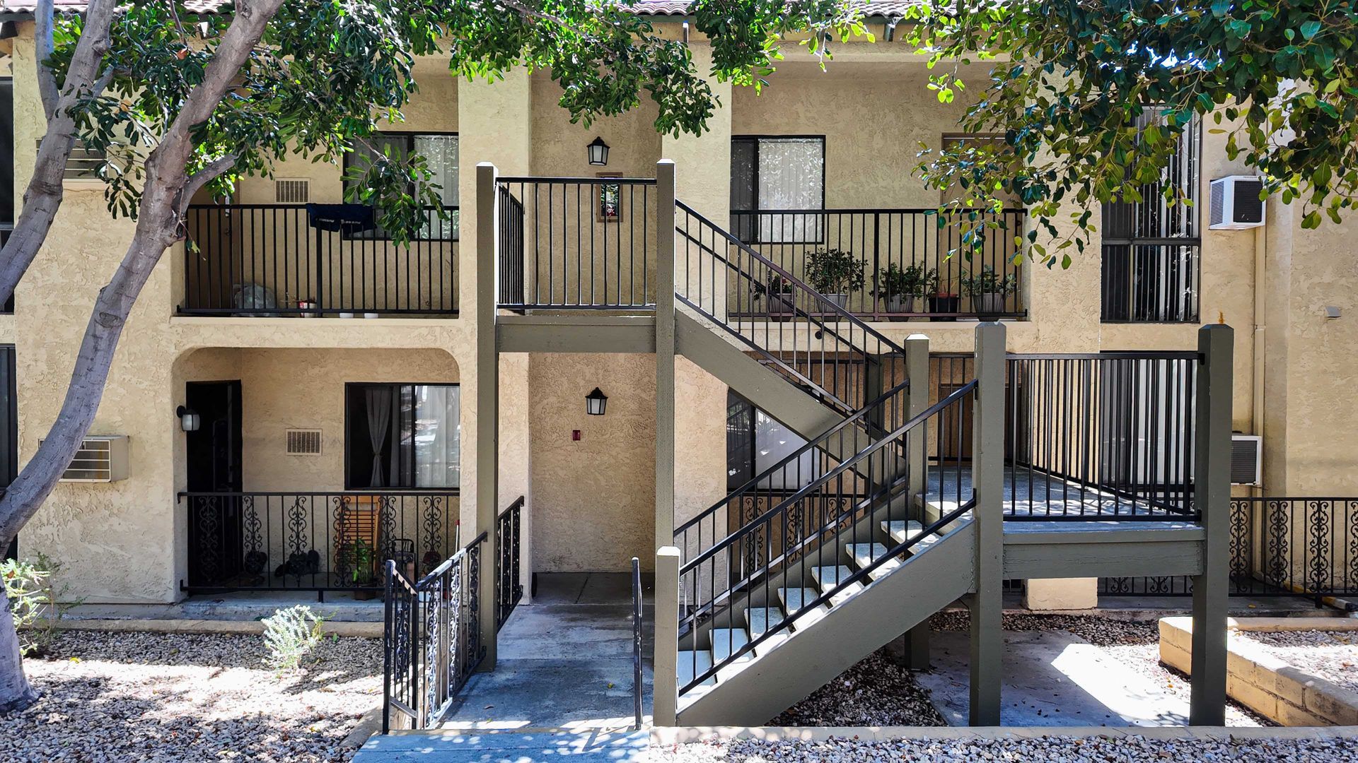 Two-story apartment building with outdoor staircases and balconies, beige exterior, trees in front.