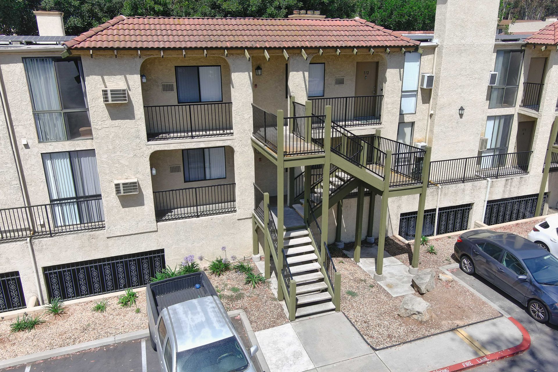 Apartment building exterior with stucco walls, balconies, stairs, and parked cars. Sunny day.