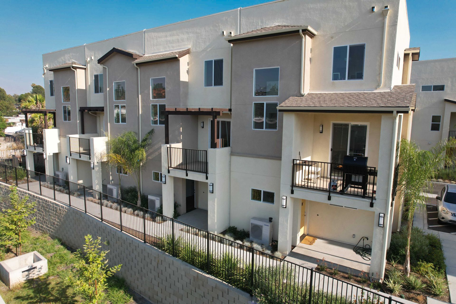 Three-story tan and gray townhouses with balconies, windows, and a fence alongside a sloped landscape.