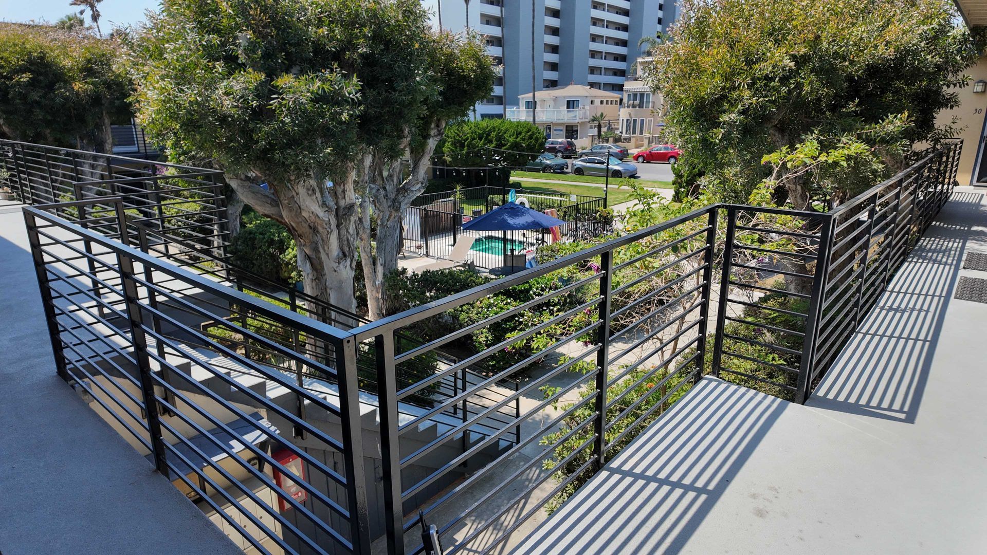 Black metal railing overlooking a courtyard with trees and a playground on a sunny day.
