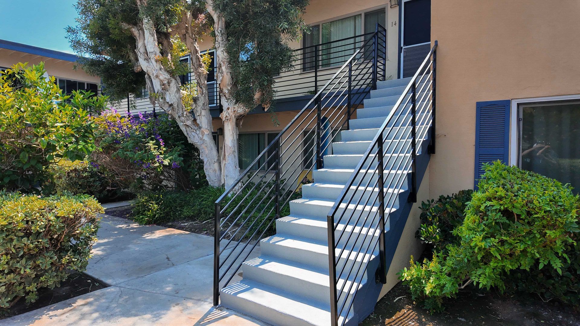 Staircase leading up to a building entrance. Features black metal railings and light-colored concrete steps. Exterior setting with greenery.