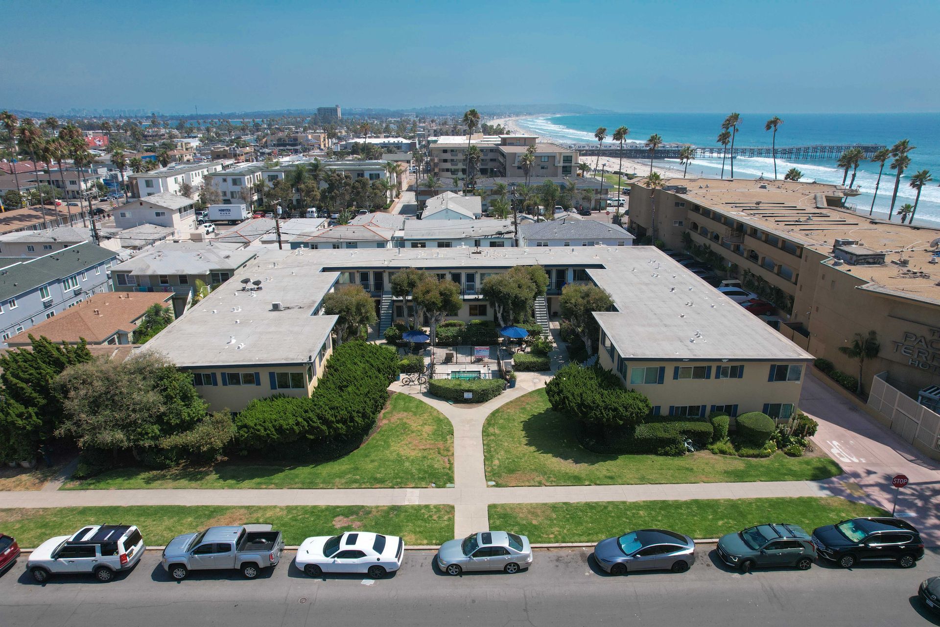 Apartment complex near beach, palm trees, parked cars, blue sky.