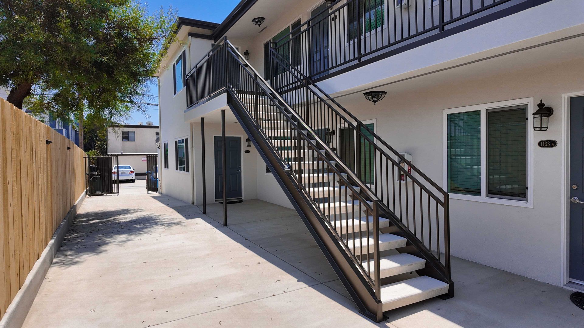 Exterior view of a two-story apartment building with an exterior staircase. Concrete pathway and wooden fence.