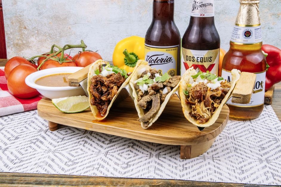 Three tacos are sitting on a wooden tray next to two bottles of beer.