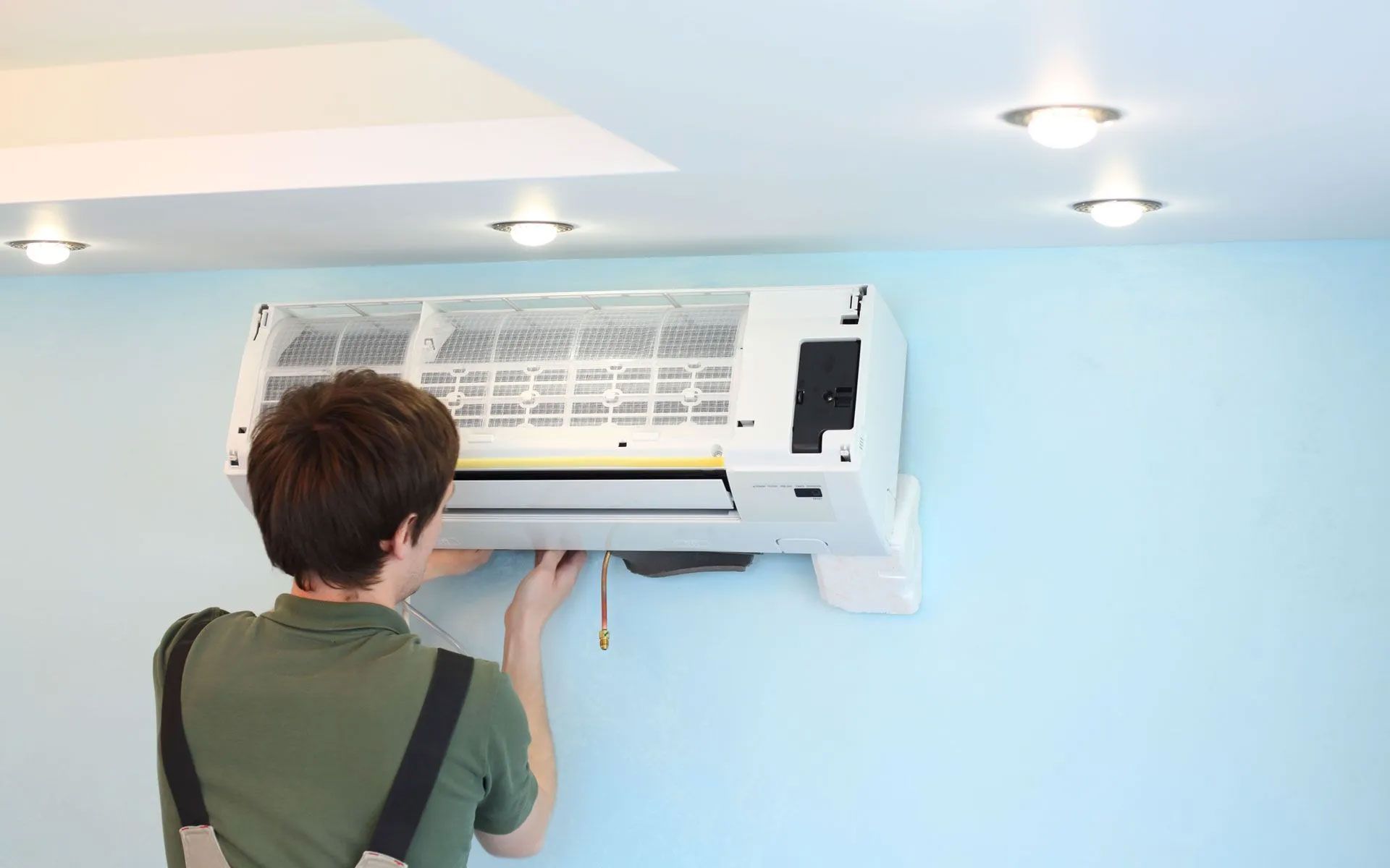 A technician wearing green work clothes installs or repairs a white wall-mounted air conditioning unit in a room.