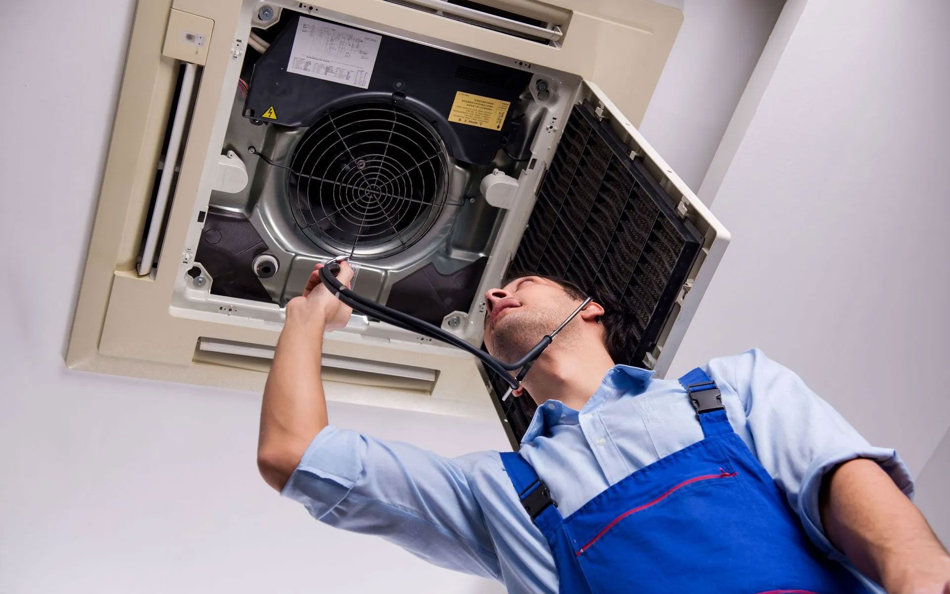 A maintenance worker in a blue uniform services a ceiling-mounted air conditioning unit.