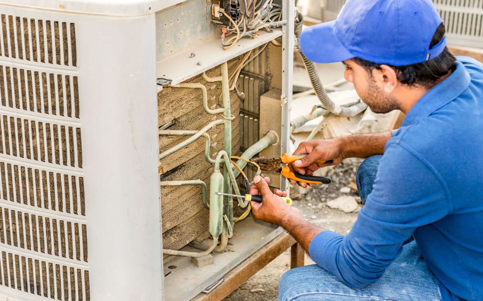A technician wearing a blue uniform and cap repairs an outdoor air conditioning unit with pliers and a screwdriver.