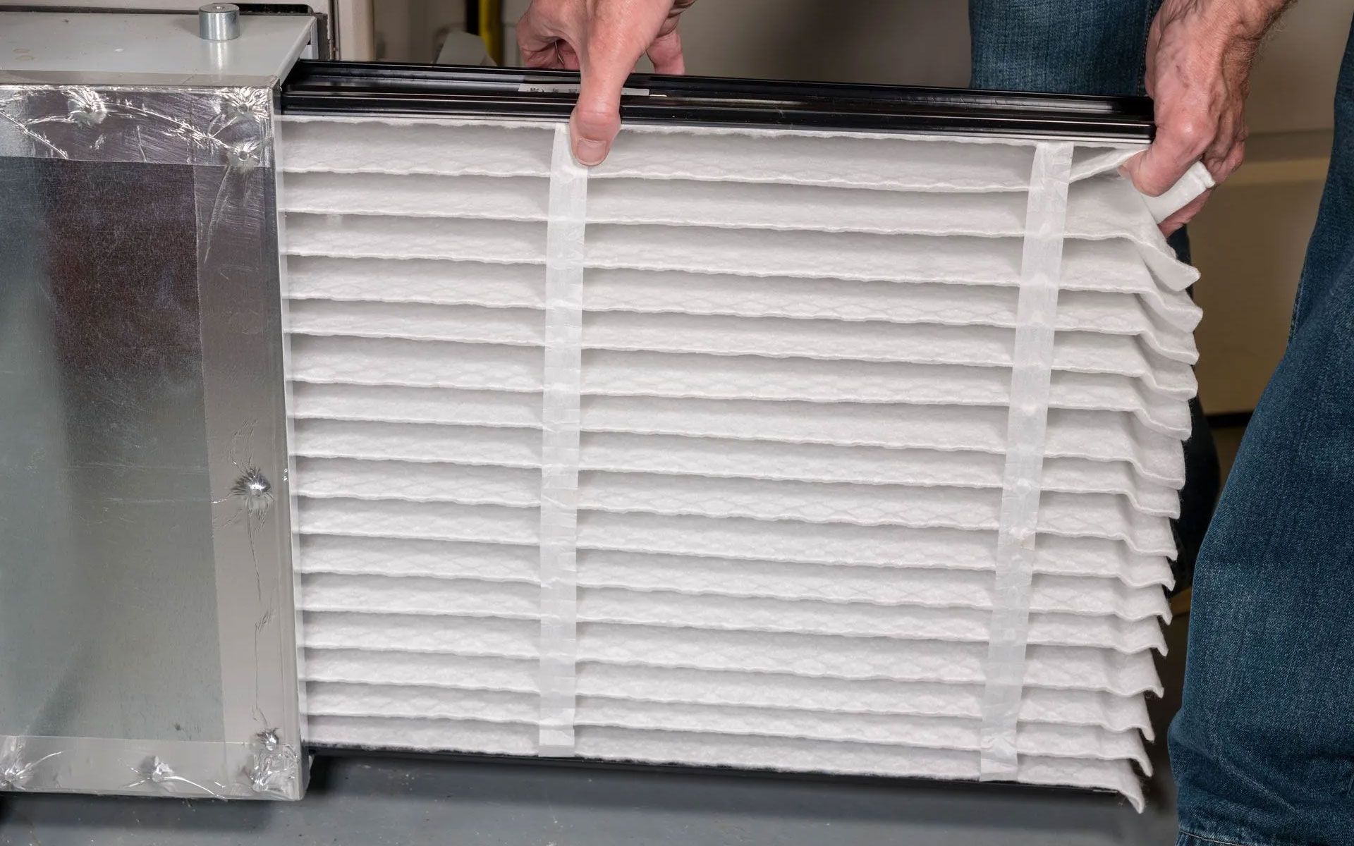 A pair of hands sliding a clean, pleated white air filter into a metal HVAC unit duct.