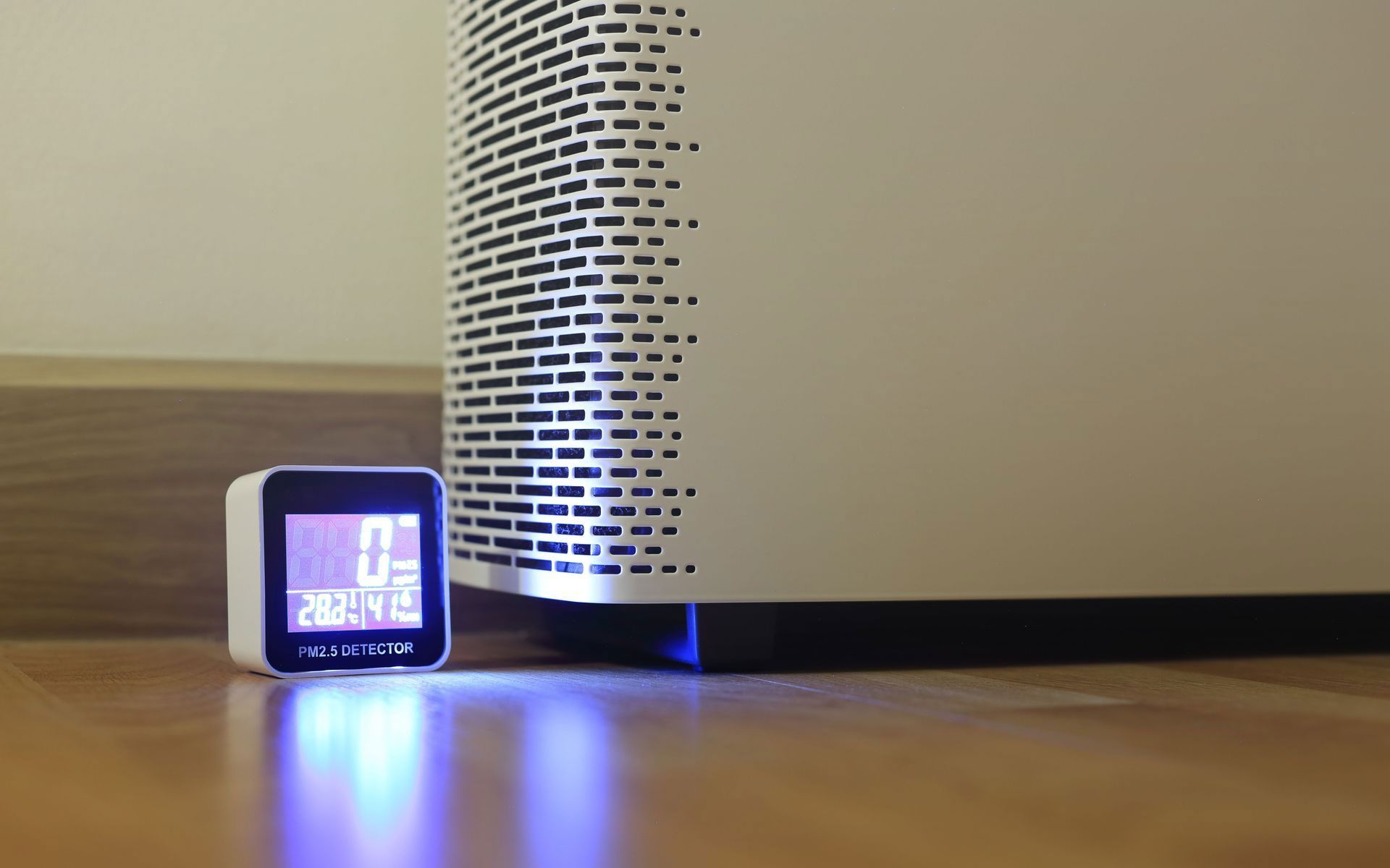 A small digital air quality monitor sits on a wooden floor next to a white air purifier with a glowing blue base.