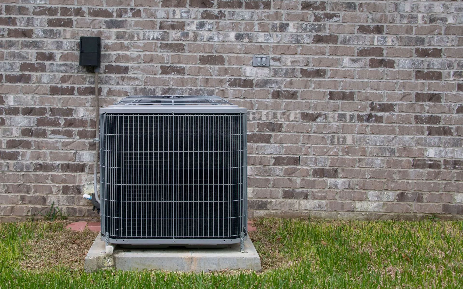 An outdoor grey air conditioning unit sits on a concrete pad against a brown brick wall above green grass.