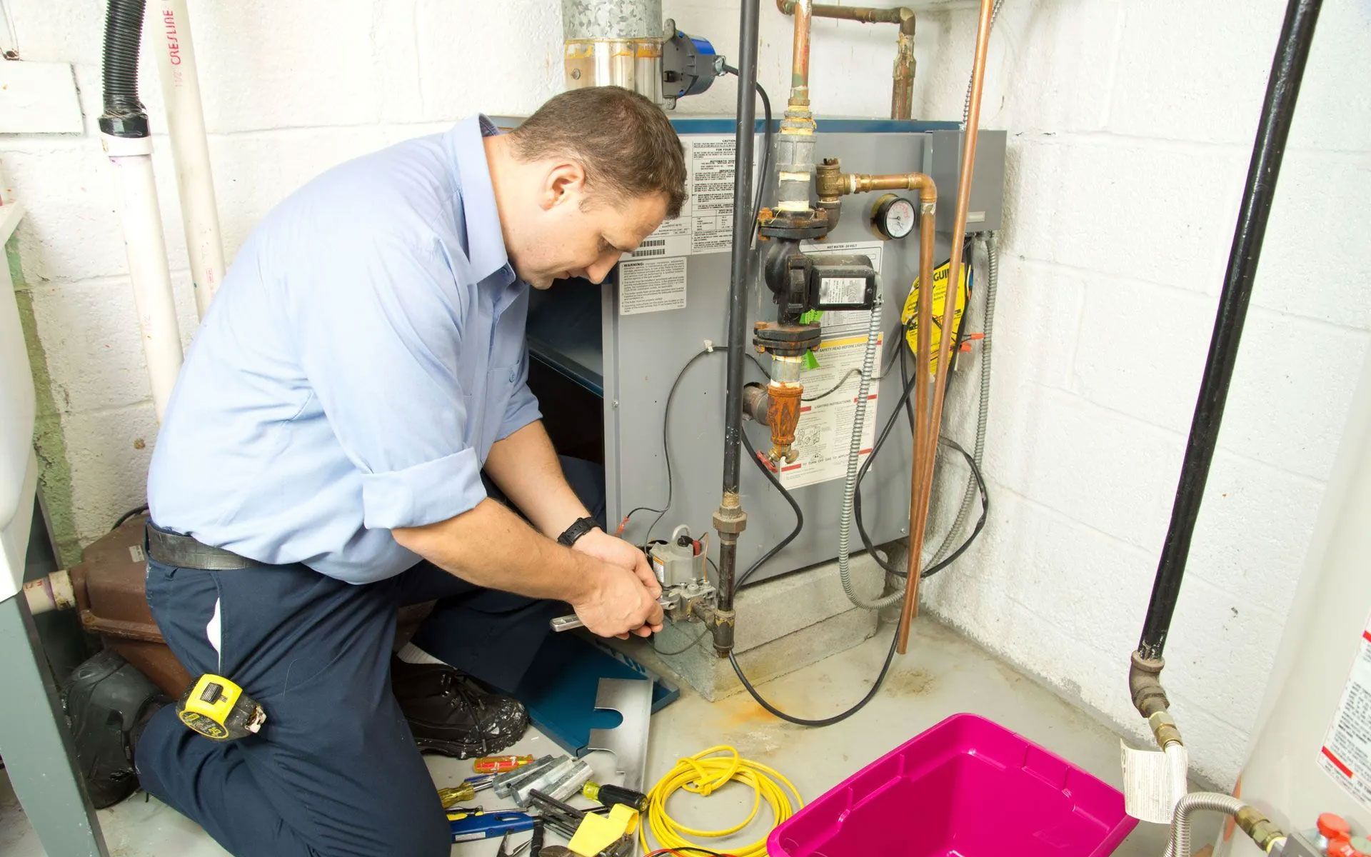 A technician in a light blue shirt works on the plumbing of a furnace in a basement, with tools and a pink bin nearby.
