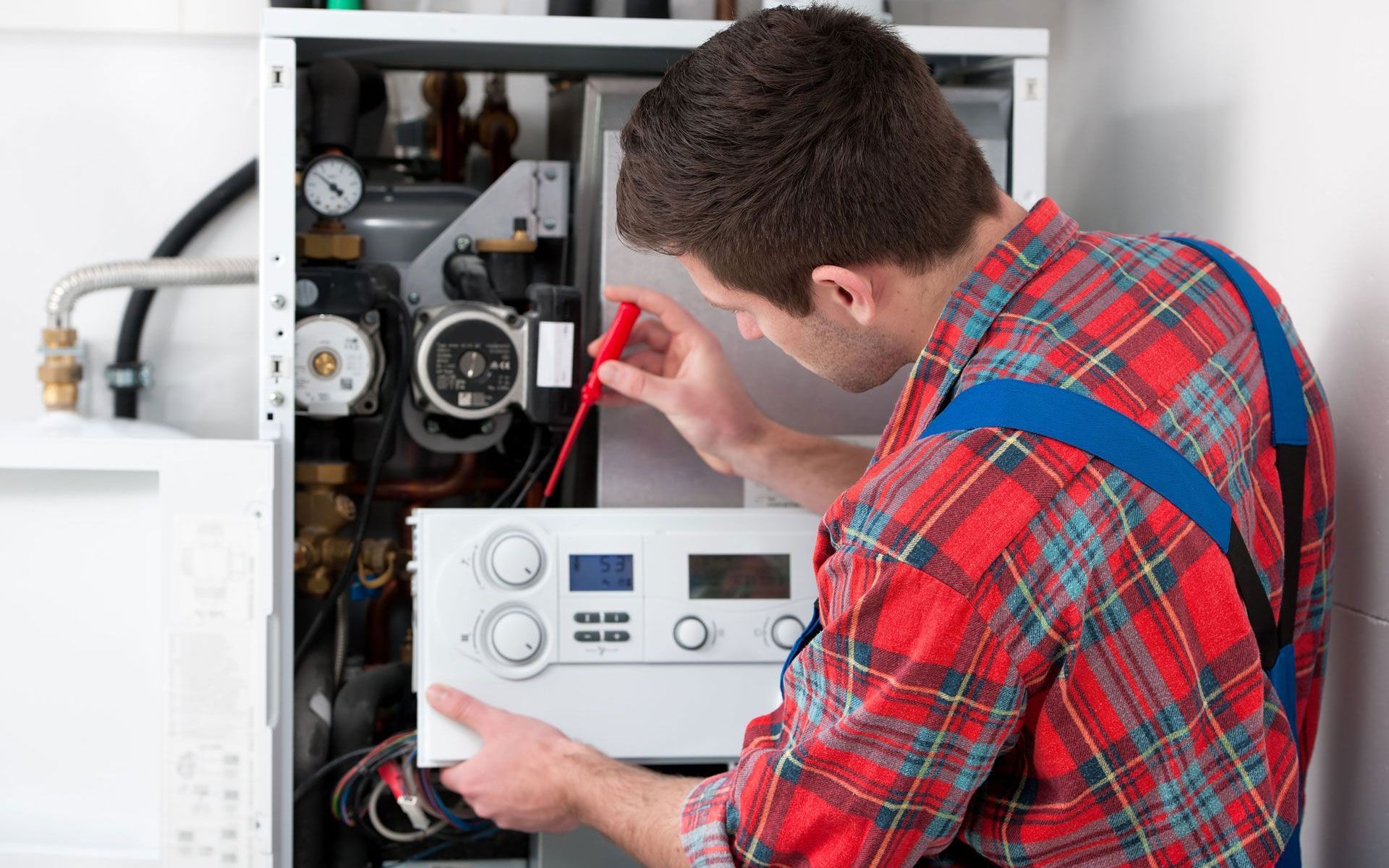 A technician in a red plaid shirt and blue overalls repairs a residential boiler, holding a red-handled tool.