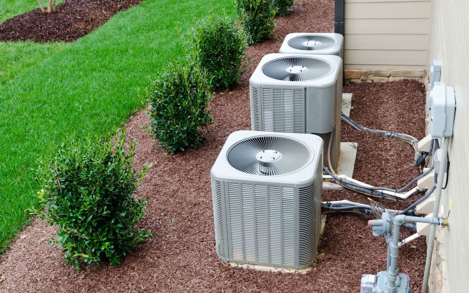 Three air conditioning units arranged in a line on a mulch bed next to a house wall and manicured lawn.