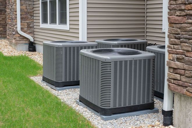 Three gray outdoor air conditioning units sit on a gravel bed next to a house with beige siding and stone trim.
