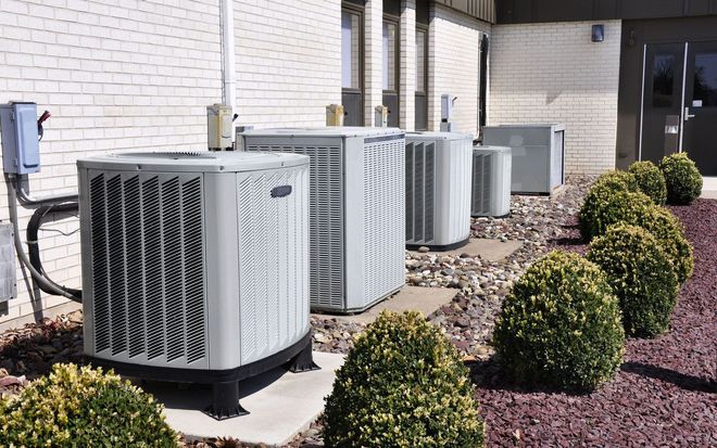 A row of light-gray industrial HVAC condenser units sits outside a white brick building next to small, rounded shrubs.