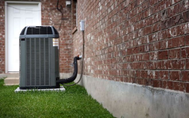 An outdoor HVAC condenser unit sits on a concrete pad next to a red brick house wall.