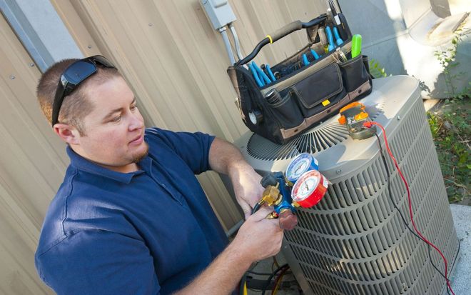 A technician services an outdoor air conditioning unit with gauges and a tool bag nearby.